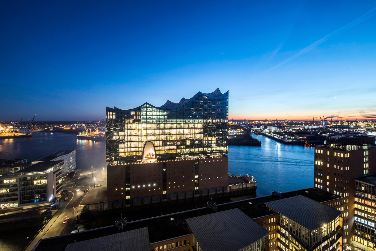 The Elbphilharmonie at blue hour from a high viewpoint, lit gold, the harbour water deep blue and the city lights behind