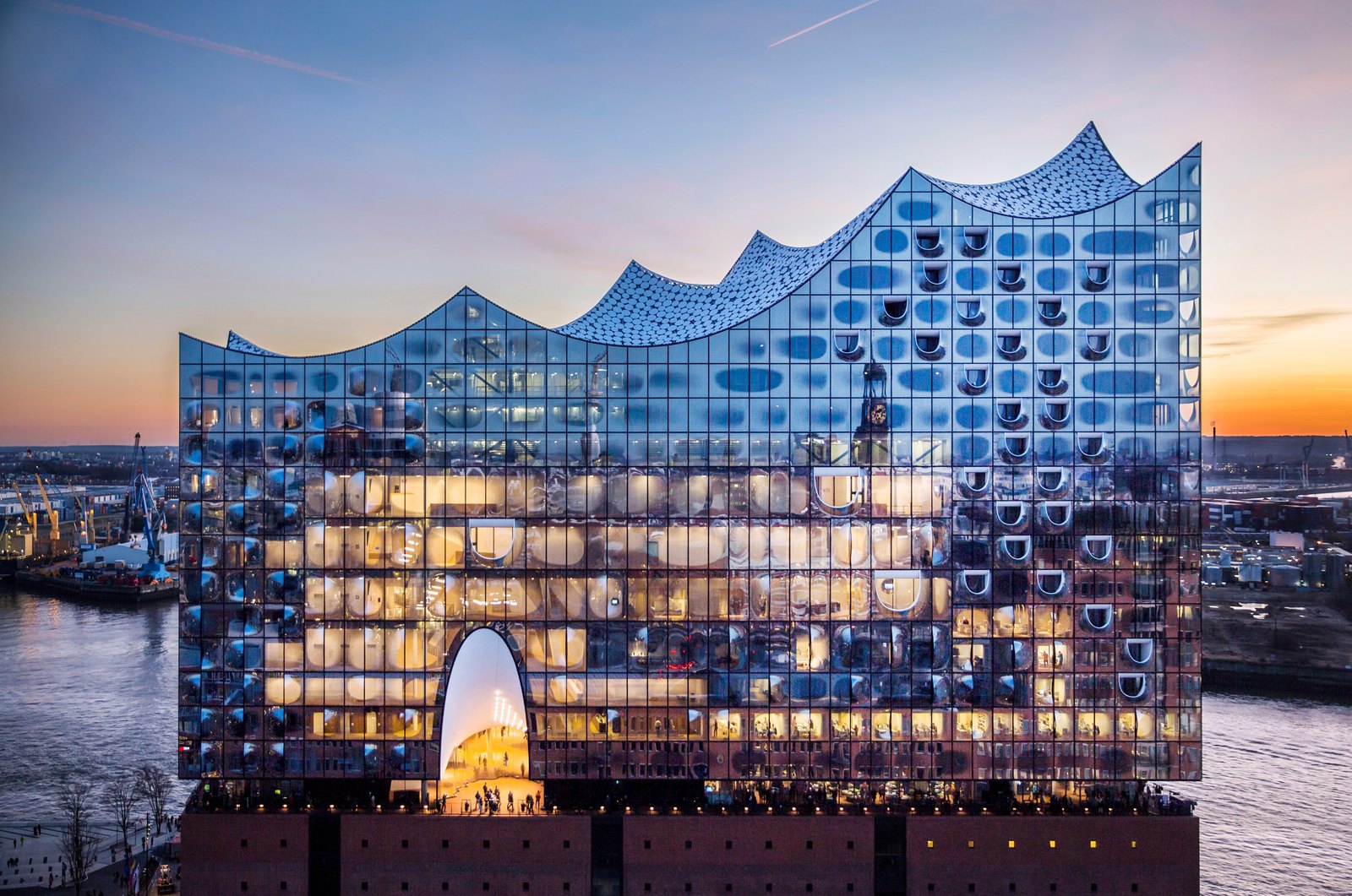 The Elbphilharmonie at dusk seen from the front, its glass upper storeys lit from within above the brick warehouse base, the arched Plaza window glowing under an orange sky
