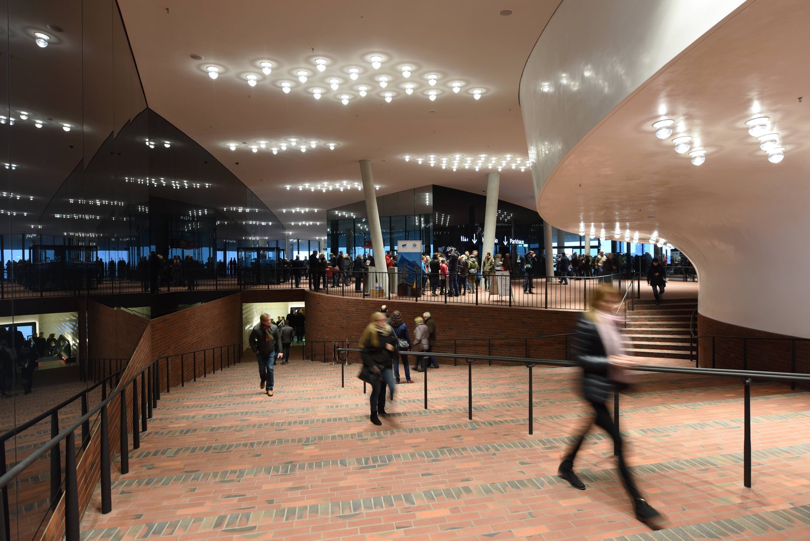 The Plaza inside the Elbphilharmonie: a brick-paved floor, a glass wall onto the harbour and rows of round ceiling lights, a visitor walking past in a blur