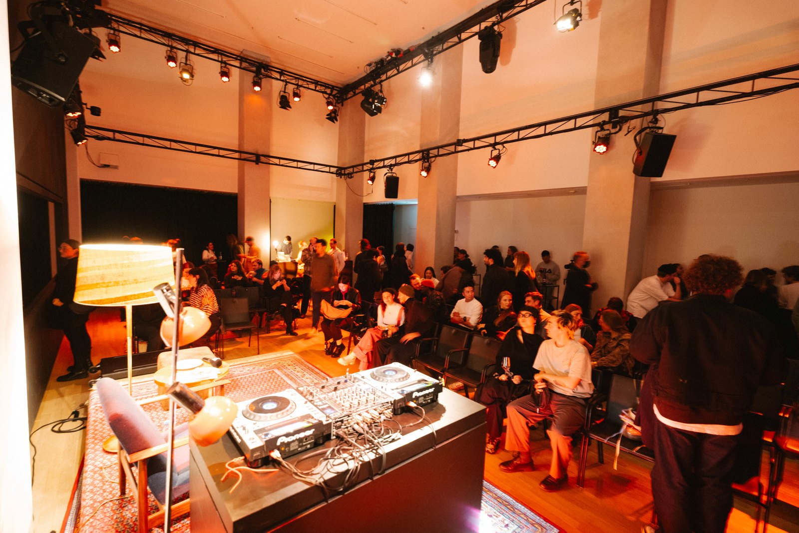 A DJ set-up with turntables and a lamp in the foreground, facing a seated audience in a warm orange room under stage lights