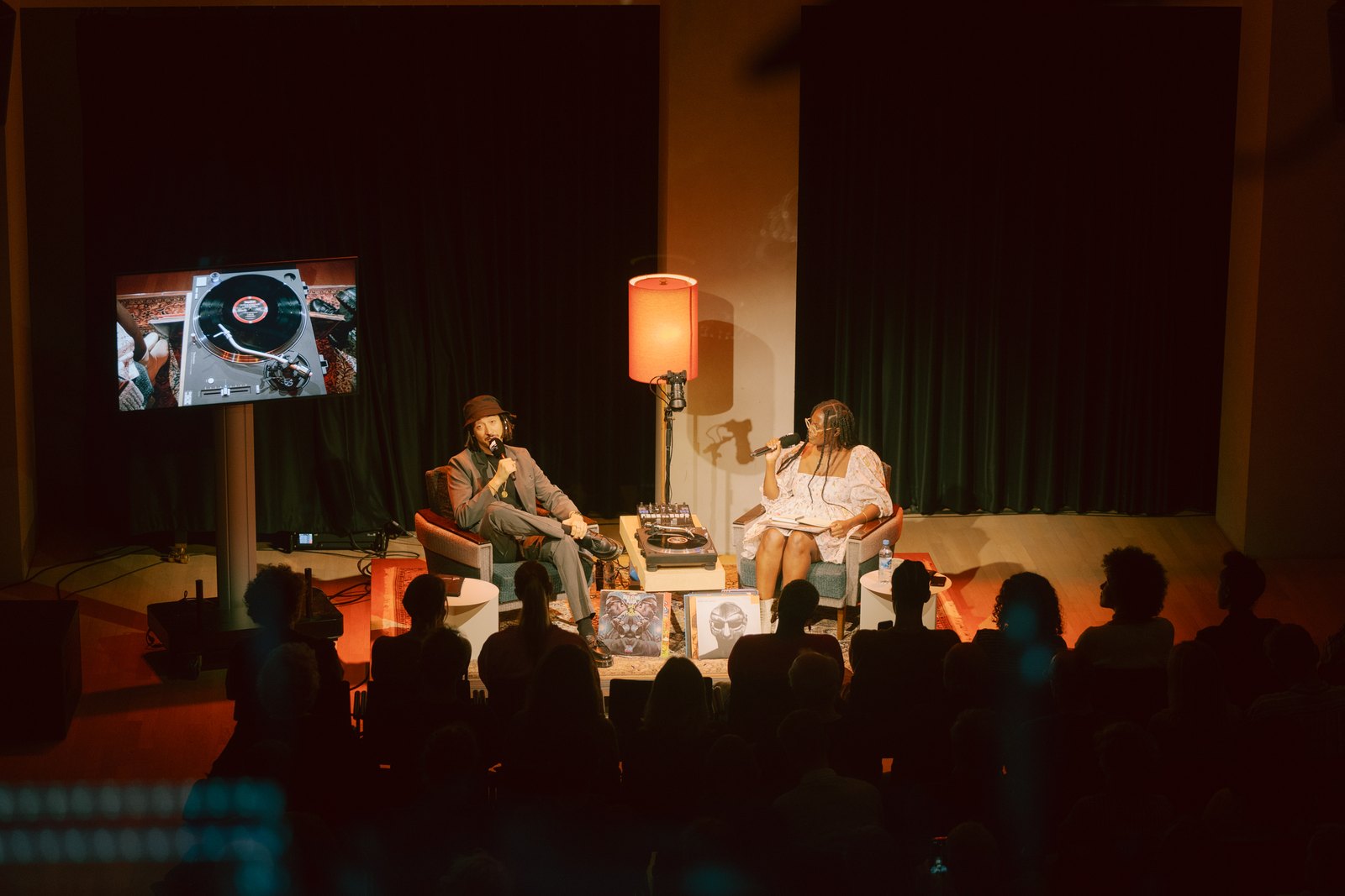 Two guests in armchairs on a lit stage beside turntables, a record shown on a screen above them, the audience in silhouette in front