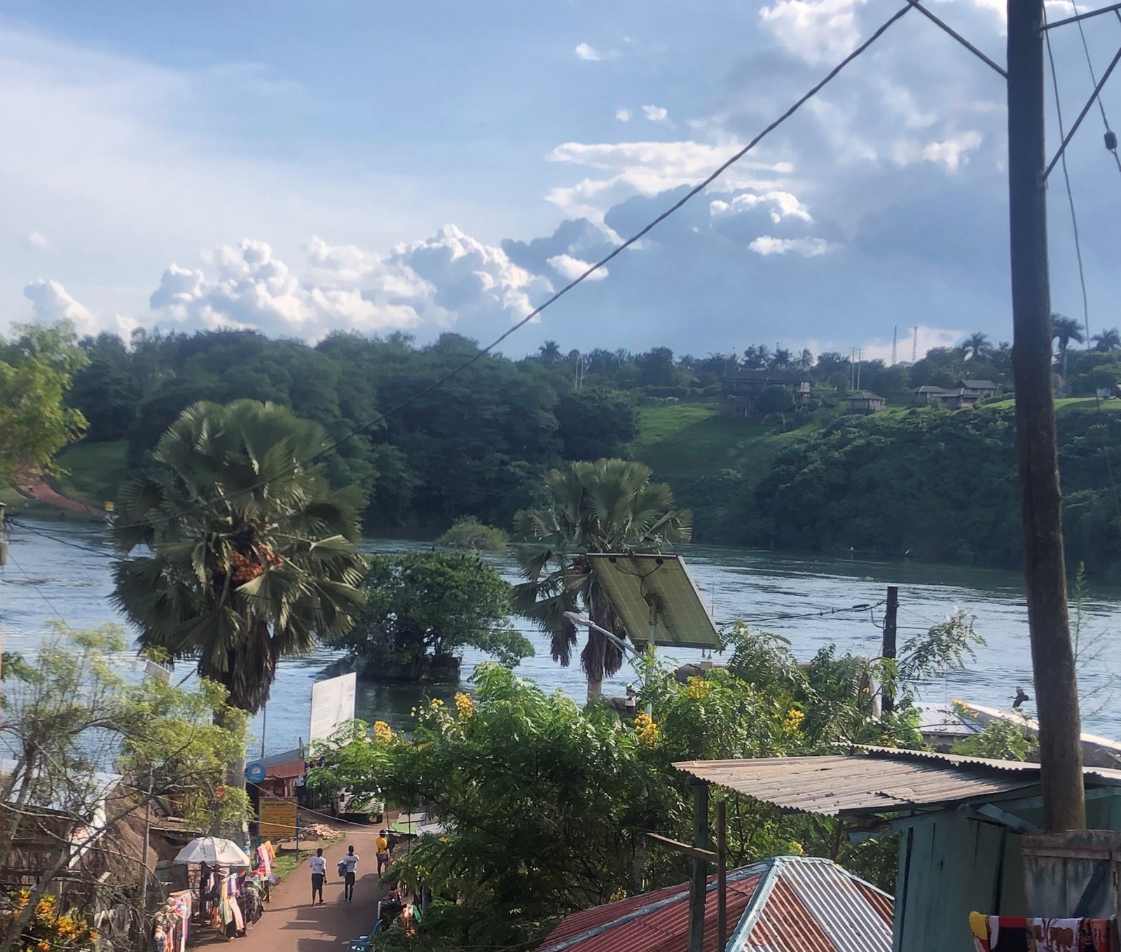 The Nile at Jinja seen from a rooftop: the wide river, palm trees and tin roofs under a big blue sky with white cloud