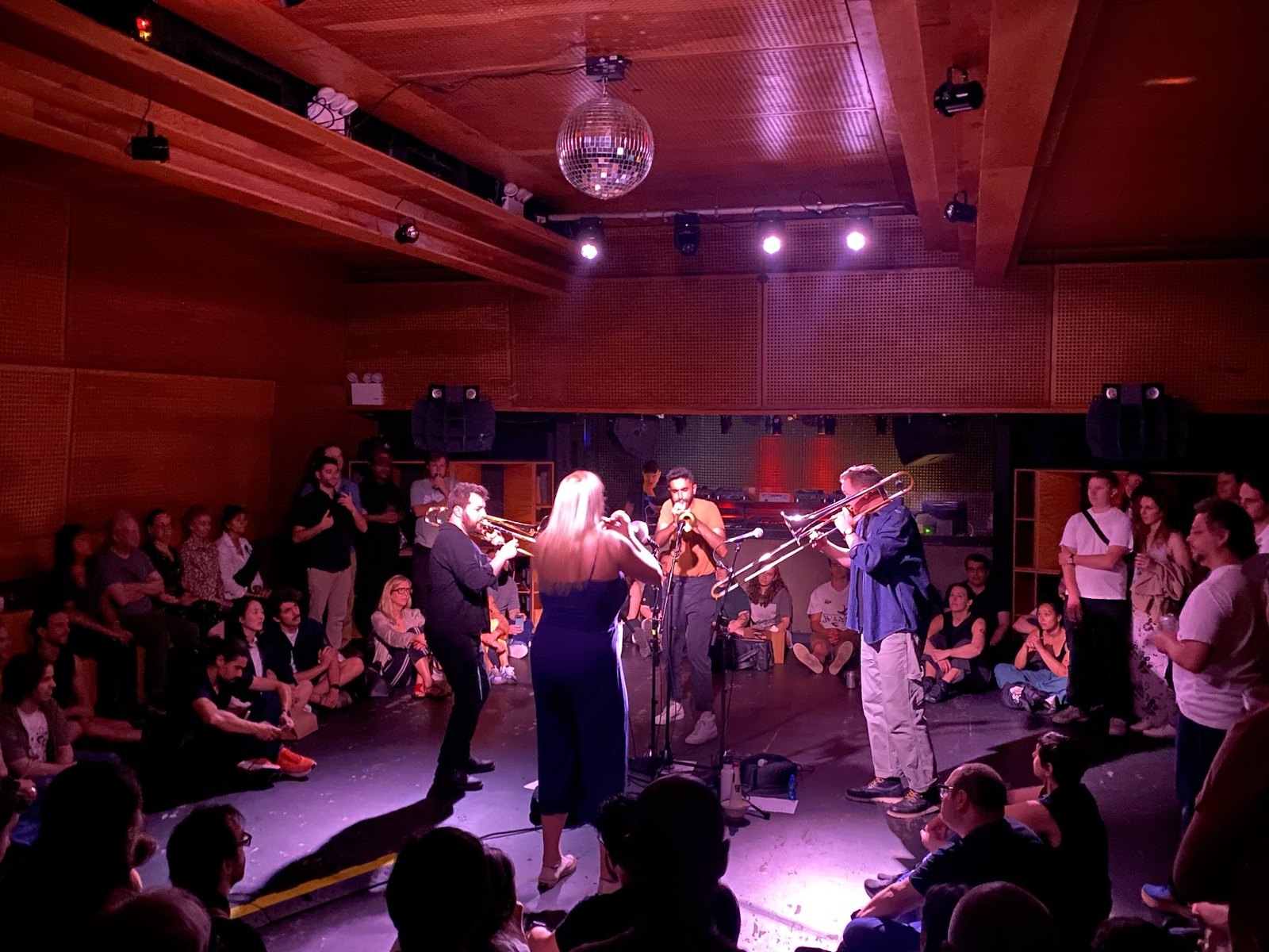A brass quartet playing in a wood-panelled room lit pink and purple, the audience sitting close around them, some on the floor