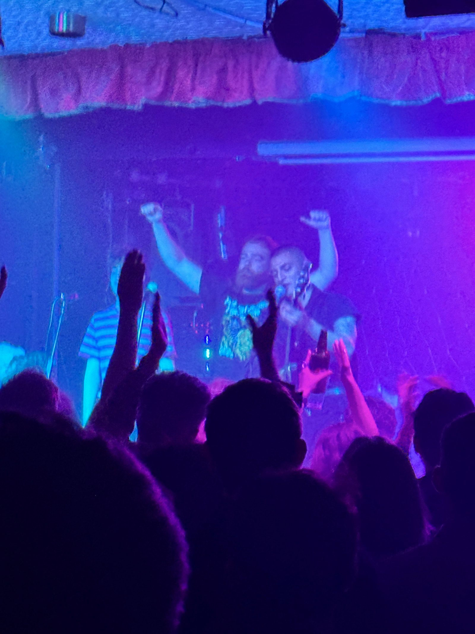 Close view of two band members with arms raised on stage, bathed in blue and violet light