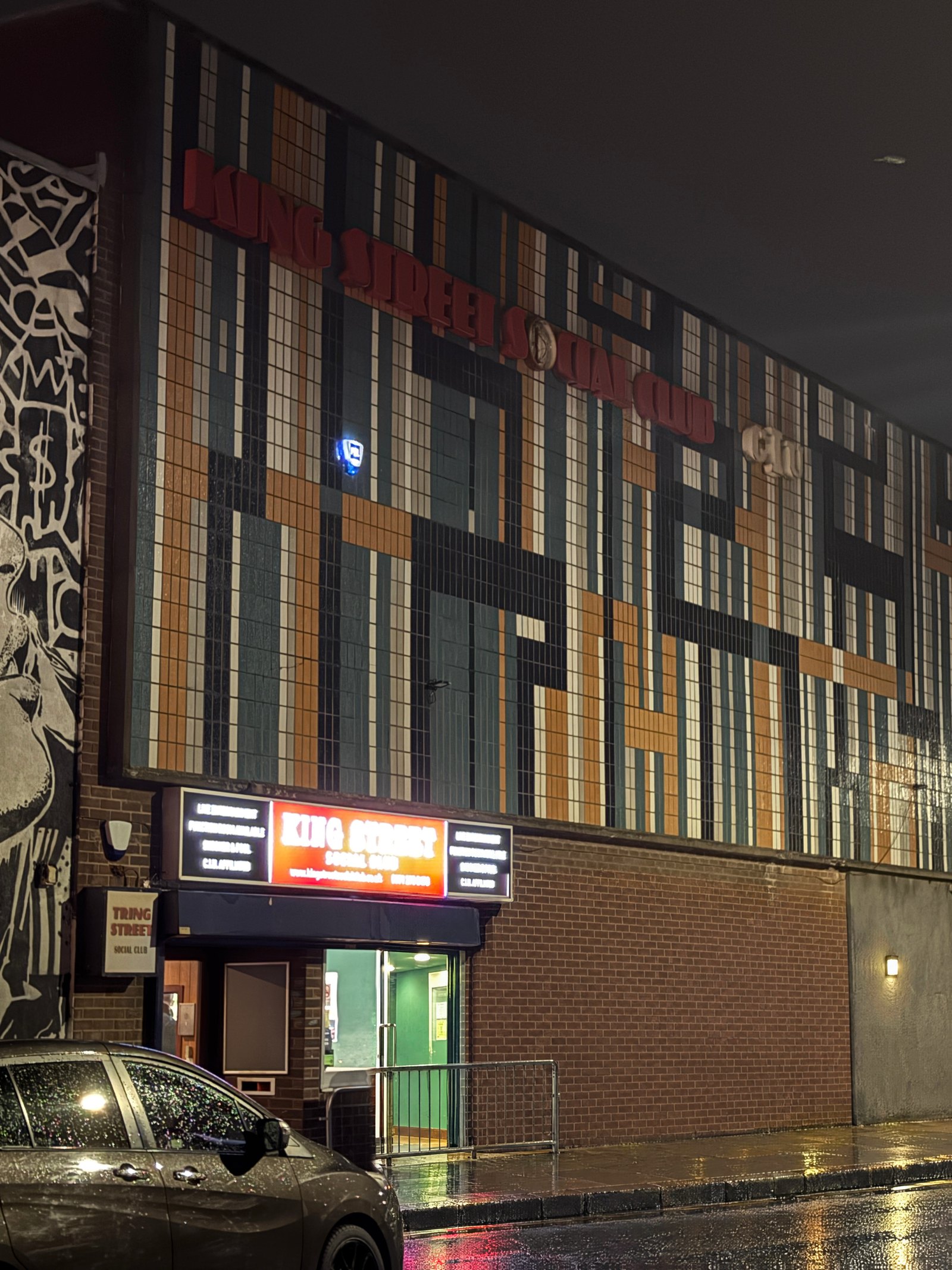Low, rain-soaked street view of King Street Social Club's tiled mosaic facade at night, red tail-light reflections on the wet tarmac, parked cars and a mural to the left