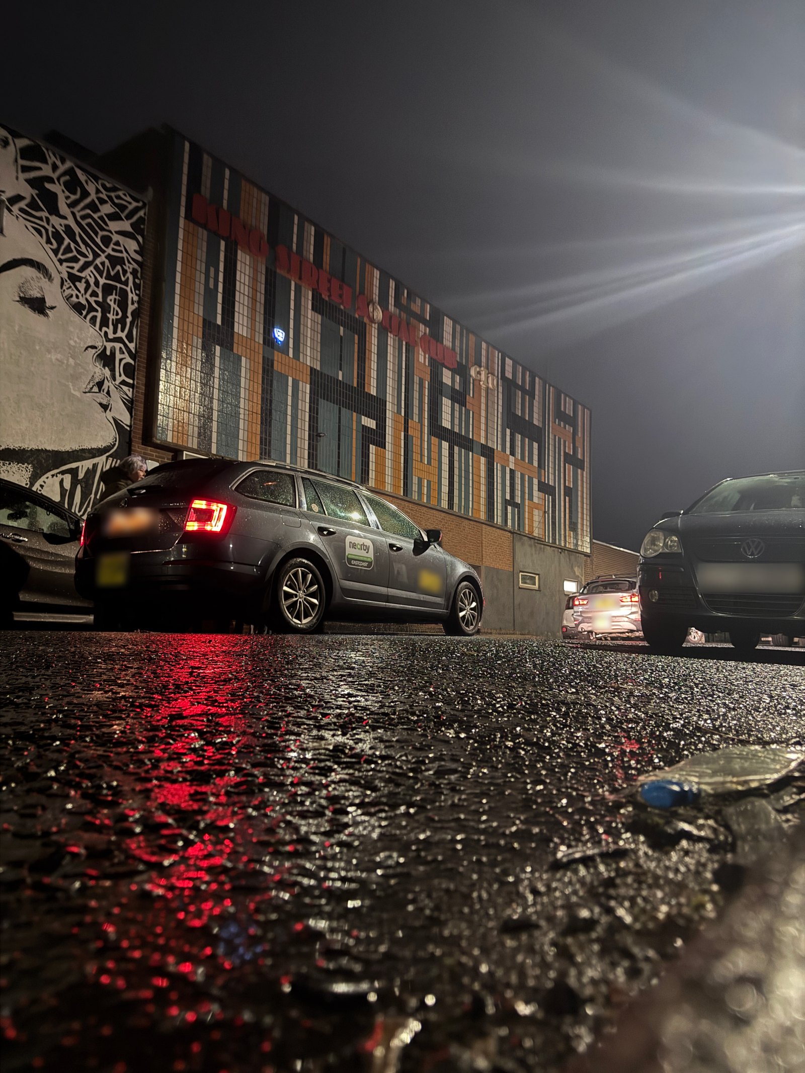 Low, rain-soaked street view of King Street Social Club's tiled mosaic facade at night, red tail-light reflections on the wet tarmac, parked cars and a mural to the left