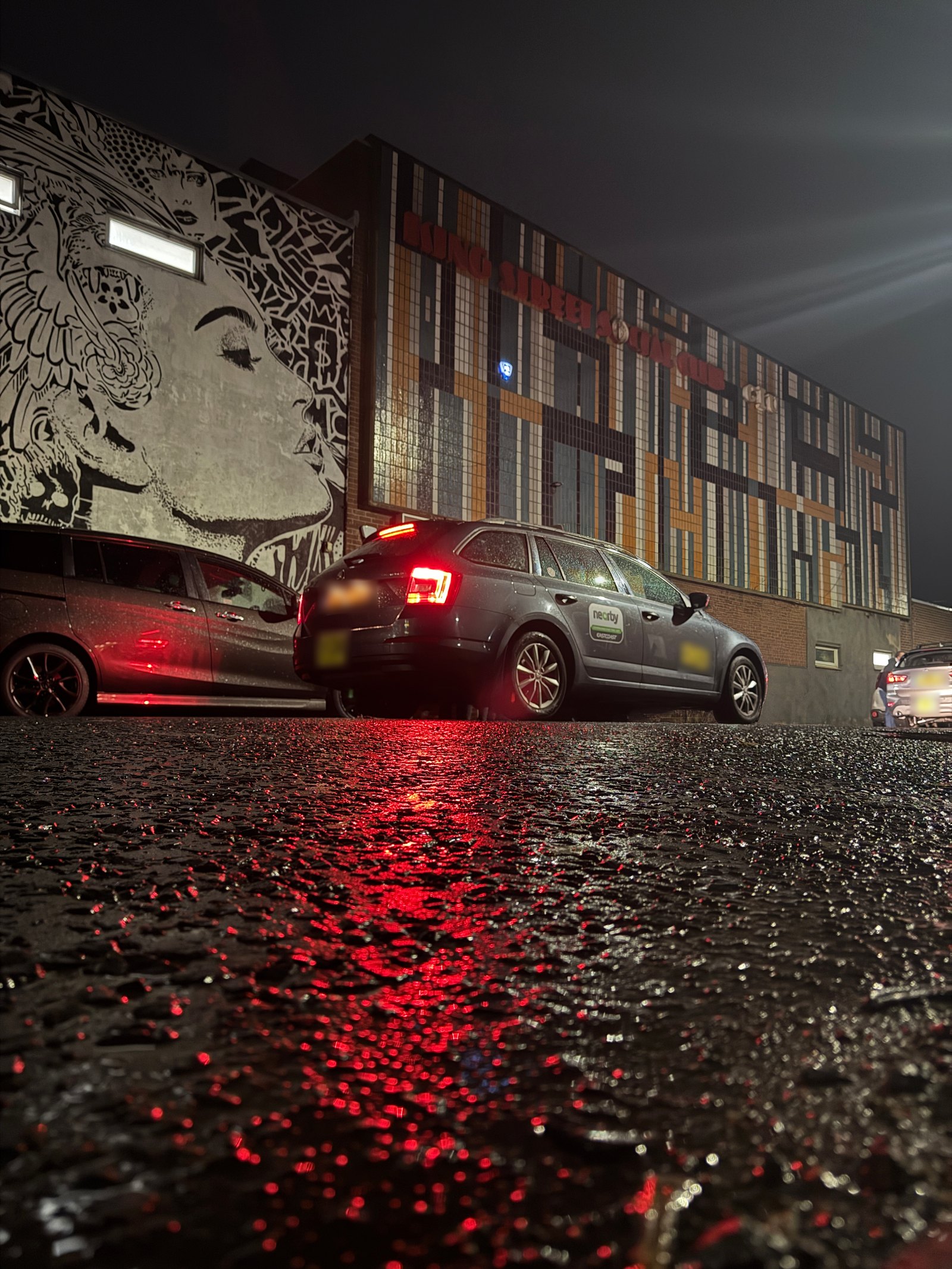 Low street view of the mural and tiled facade at night, red tail-light reflections on wet tarmac