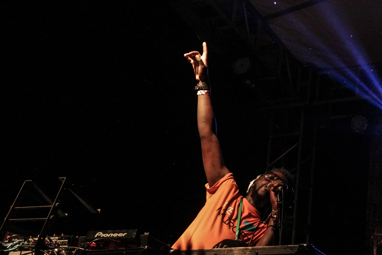 A performer on stage at Nyege Nyege with one arm raised under a blue beam of light, a mixer in front