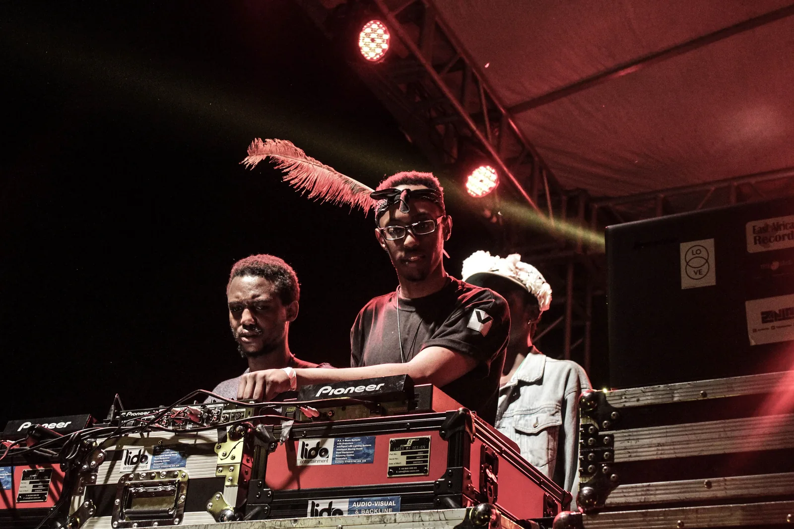 Two DJs at a mixer under red stage lights at Nyege Nyege, one wearing a feathered headdress, a crowd barrier in front