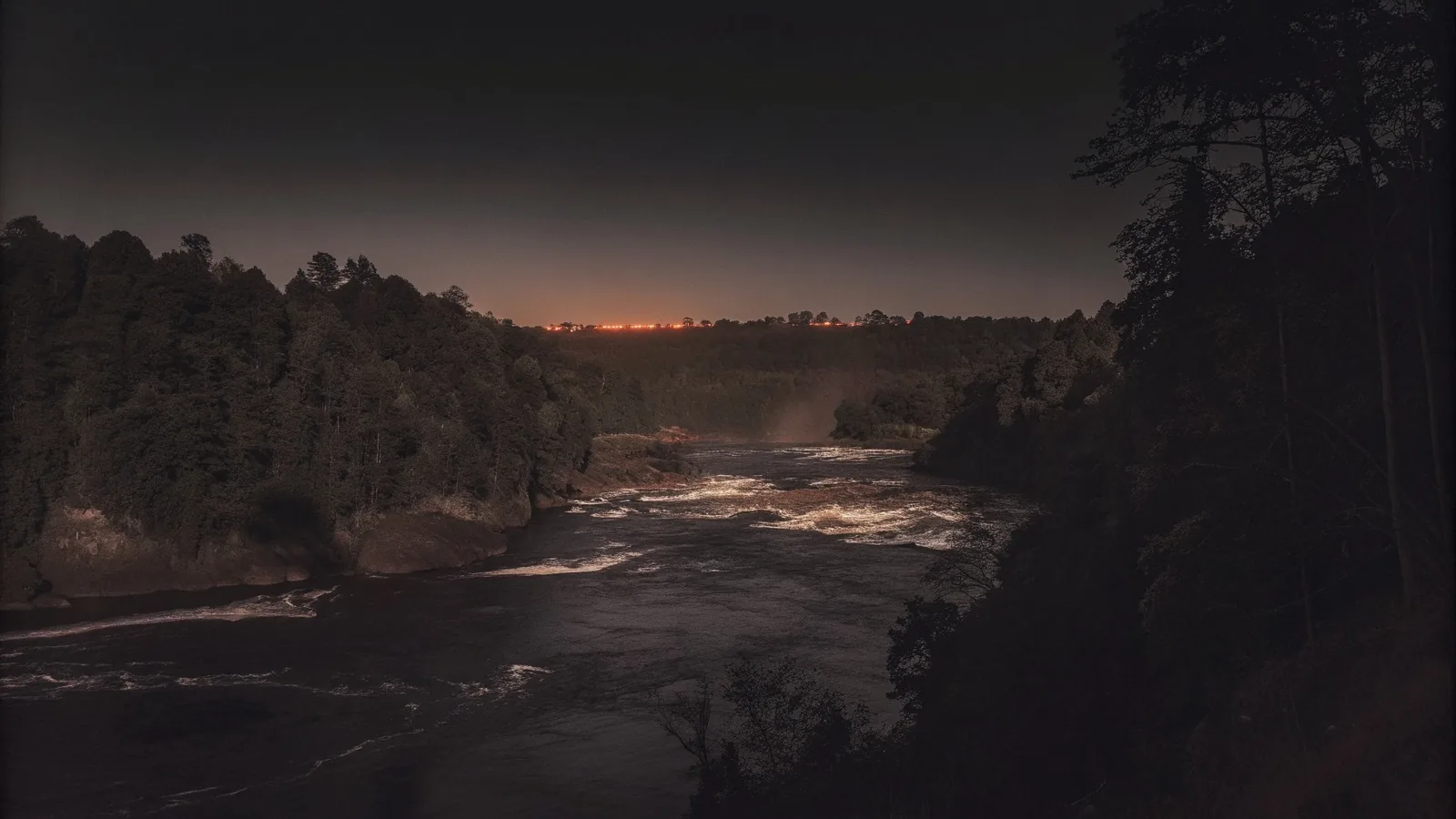 River rapids between dark wooded banks at night, a glow on the horizon