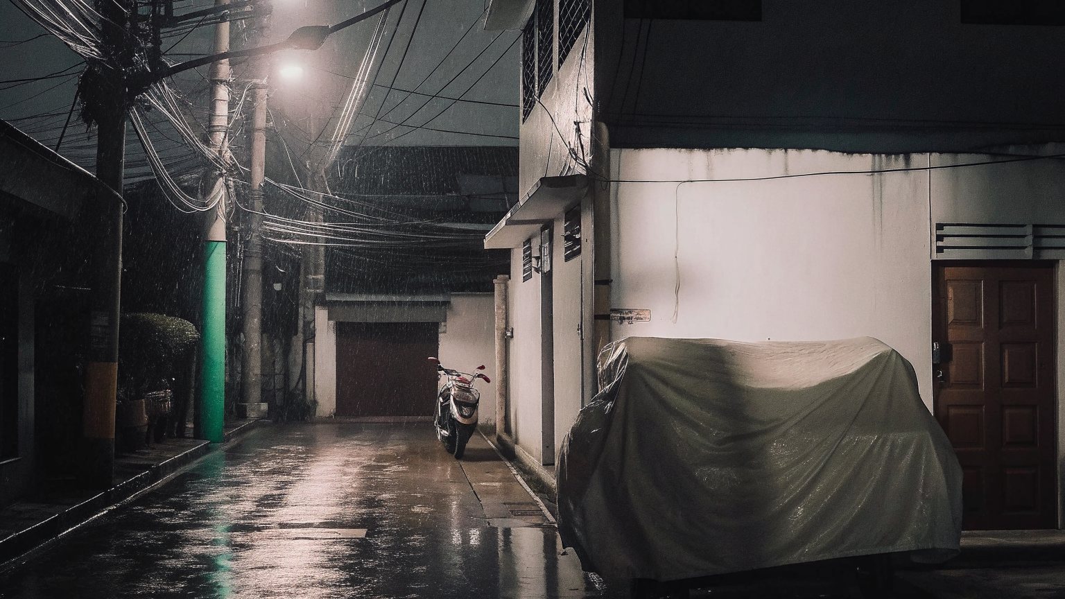 A narrow side street after rain at night, overhead wires, a covered scooter and a lit doorway