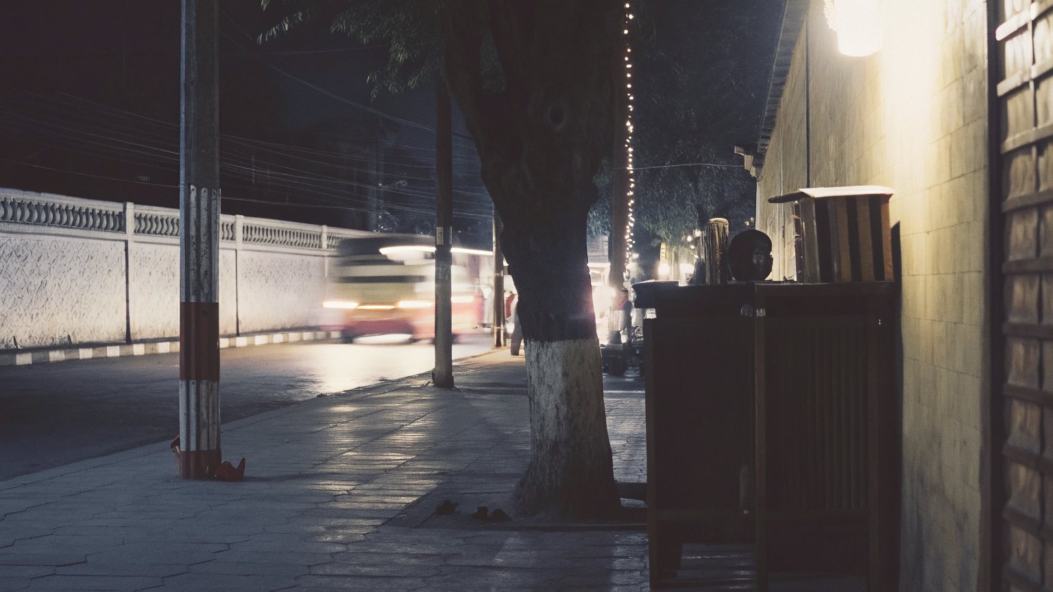A pavement at night beside a white wall and a tree, a car blurring past