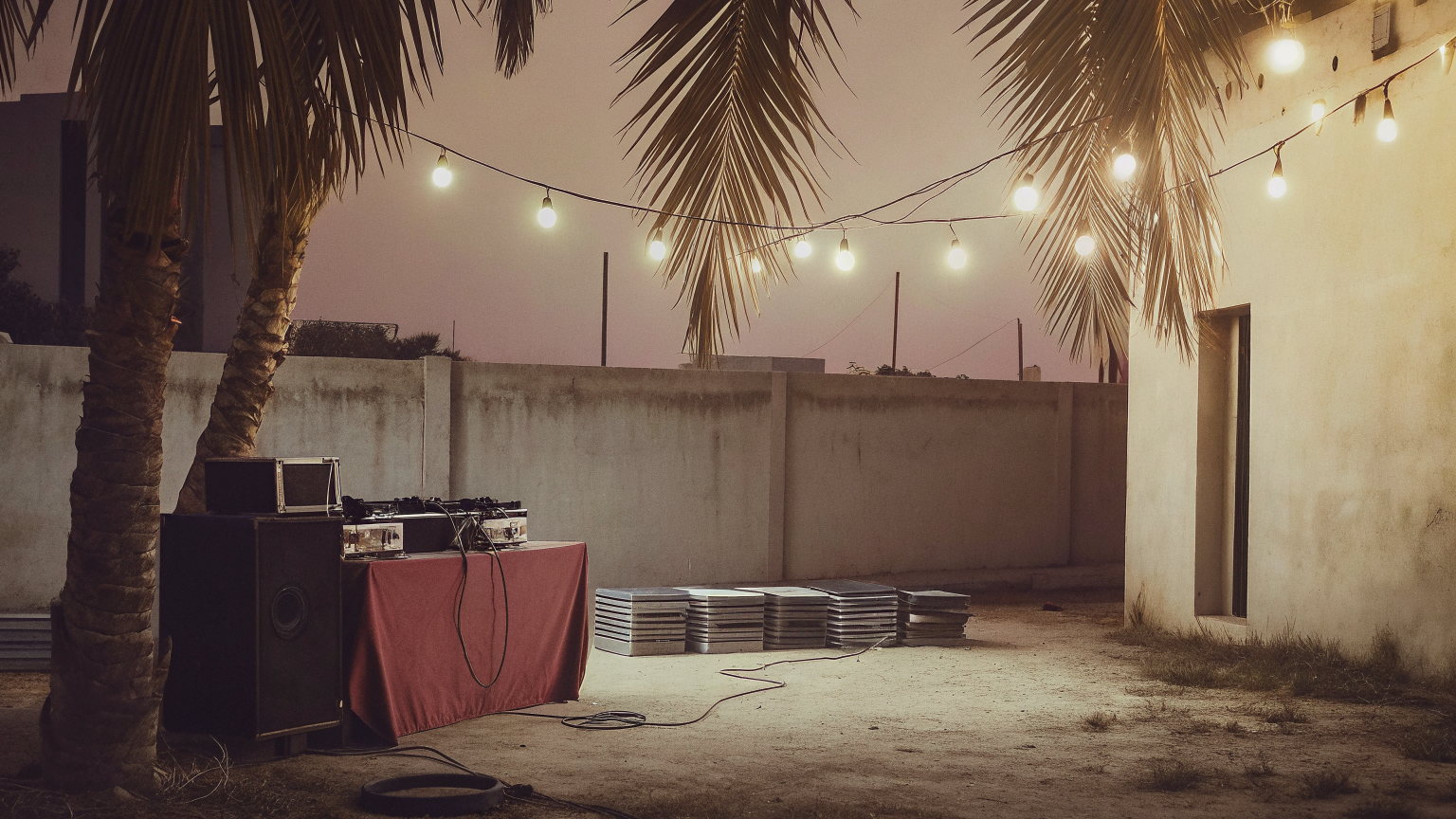 A bar garden in Osu, Accra at dusk, festoon bulbs under palms, DJ decks on a cloth-covered table and a speaker, nobody there.
