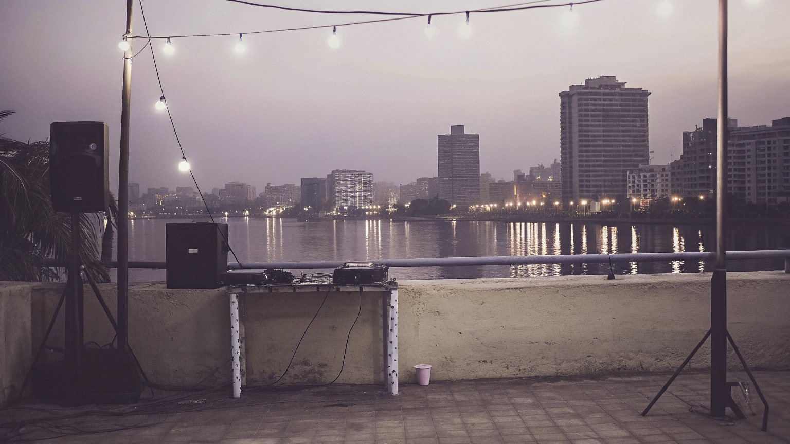 A Cairo rooftop at dusk above the Nile, festoon bulbs, two speakers and a DJ table at the parapet, the towers of the far bank reflected in the river.