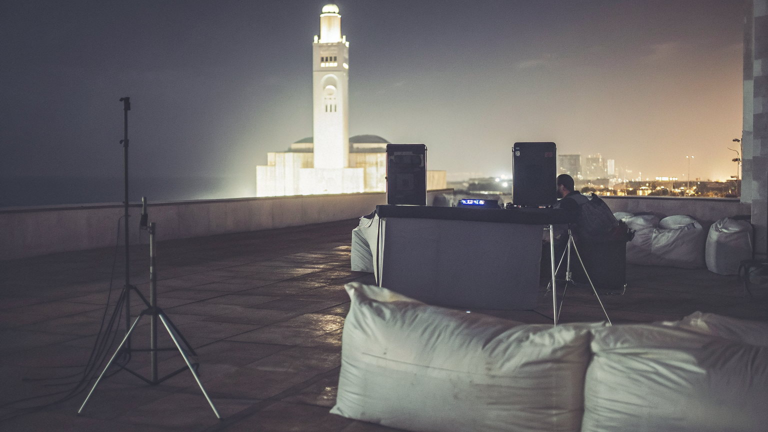 A rooftop terrace on the Casablanca Corniche at night, cushions and a DJ table, the lit minaret of the Hassan II Mosque on the shore behind.