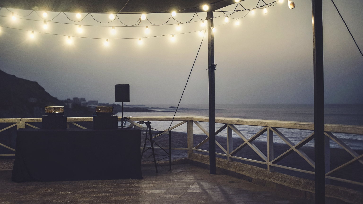 A seafront terrace on the Almadies coast of Dakar at dusk, festoon bulbs over a cloth-covered DJ table and a speaker, Atlantic surf below.