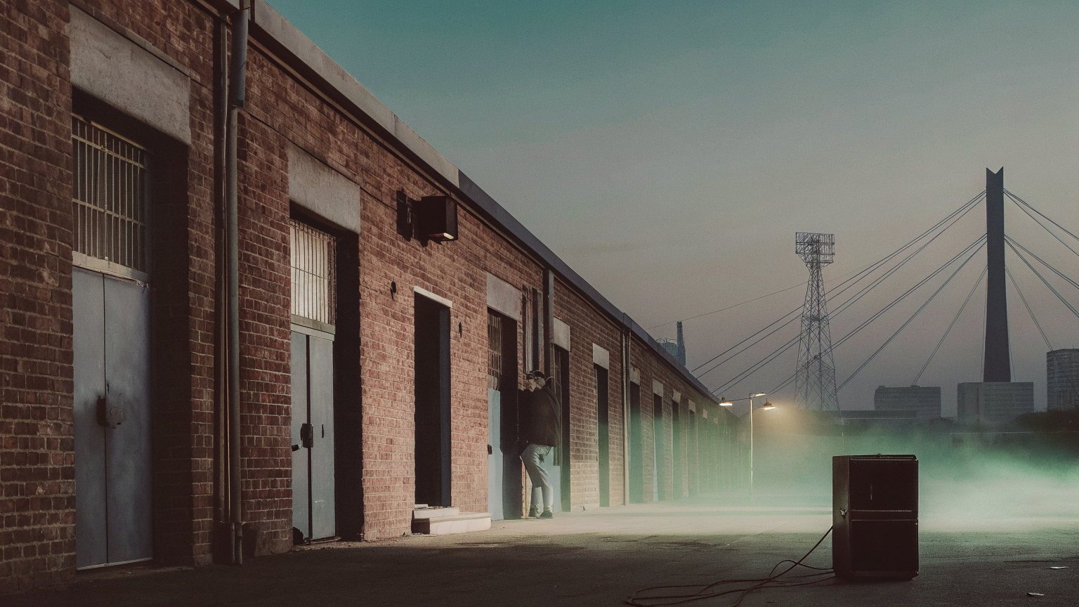 A Newtown warehouse street in Johannesburg at dusk, a speaker and cables on the road, haze, the Nelson Mandela Bridge pylon behind.