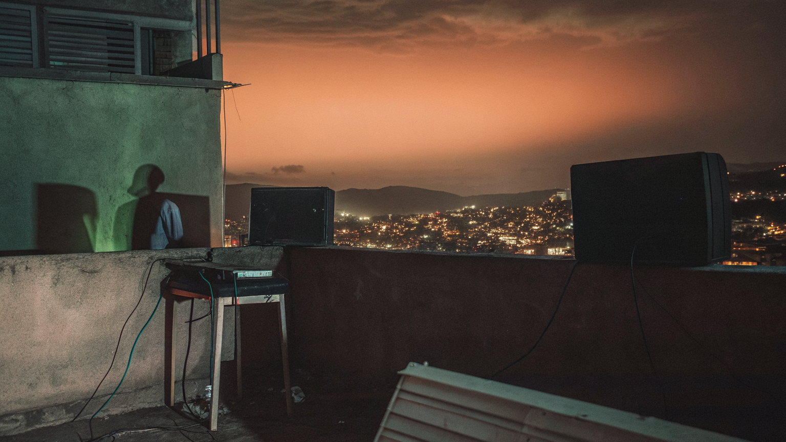 A Kampala rooftop at dusk, two speakers and a mixer on a stool, cables on the concrete, a shadow on the wall, the hills of the city lit below.
