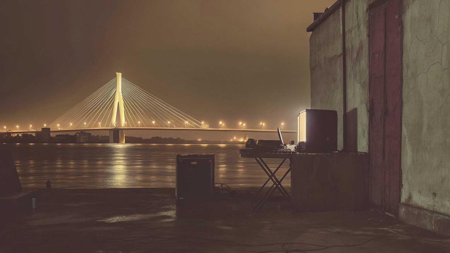 The lit Lekki-Ikoyi bridge across the Lagos lagoon at night, a speaker and a DJ table on wet concrete by a warehouse door.