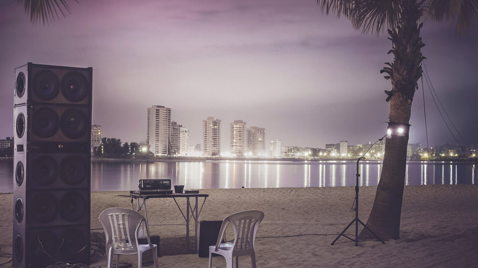 A beach on the Ilha de Luanda at dusk, a speaker stack, a DJ table and two plastic chairs on the sand, the lit Luanda skyline across the bay.